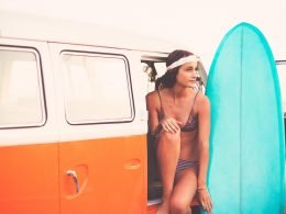 A woman sits in the open door of her camper van holding her surf board.