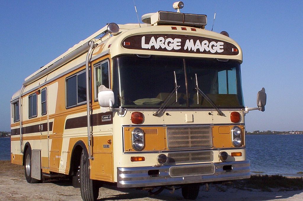 A vintage brown and yelllow BlueBird Wanderlodge name Large Marge parked near the water.